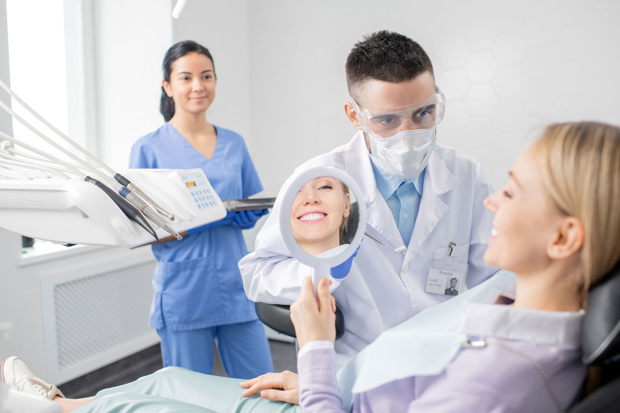 Paciente rubia sonriendo ante un espejo de mano junto a un dentista con mascarilla y gafas, con una asistente detrás