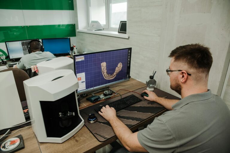 Técnico dental con gafas diseñando en el ordenador una arcada en 3D, junto a un escáner de laboratorio