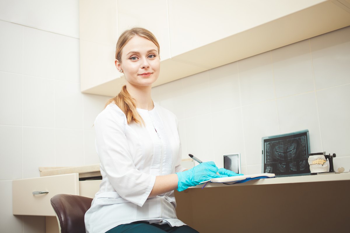 Odontóloga con bata blanca y guantes azules tomando notas en una carpeta, con una radiografía y un modelo dental al fondo