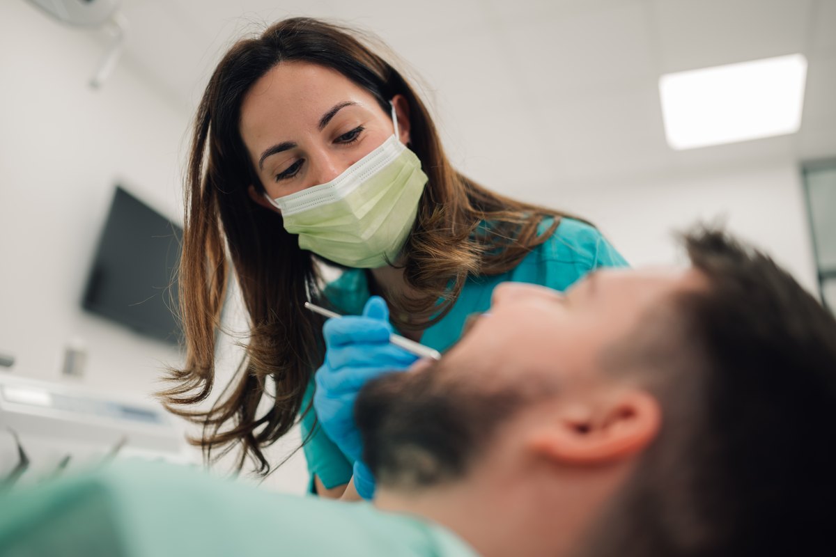 Dentista con mascarilla y guantes azules revisando la boca de un paciente con barba tumbado en el sillón
