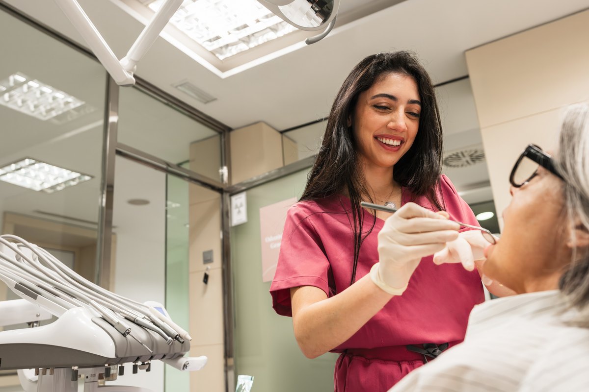 Odontóloga sonriente con uniforme granate revisando con un espejo dental a una paciente mayor con gafas