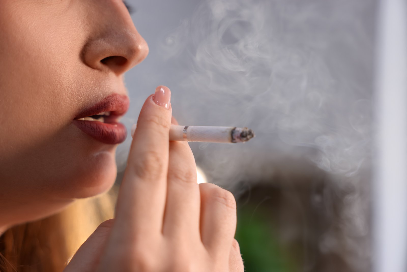Primer plano de unos labios pintados junto a un cigarrillo encendido y su humo